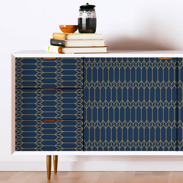 A white sideboard with drawer fronts and cabinet doors covered in navy and gold geometric peel and stick paper, with books and a lidded jar on top.