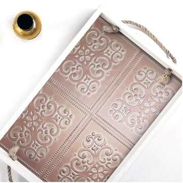 A white wooden serving tray with braided rope handles, its inside base lined with dusty rose and copper embossed tile-pattern peel and stick paper, photographed from above on a white surface beside a small gold-rimmed glass vase.