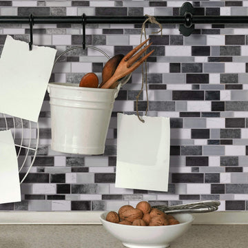 A kitchen backsplash of black, white and grey brick-shaped mosaic tiles, with a hanging rail, a white bucket of wooden utensils and a bowl of walnuts.
