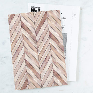 A single sheet of wood grain herringbone peel and stick wallpaper resting on its printed packaging insert, which shows a Crack and Peel Here guide, on a white marble surface.
