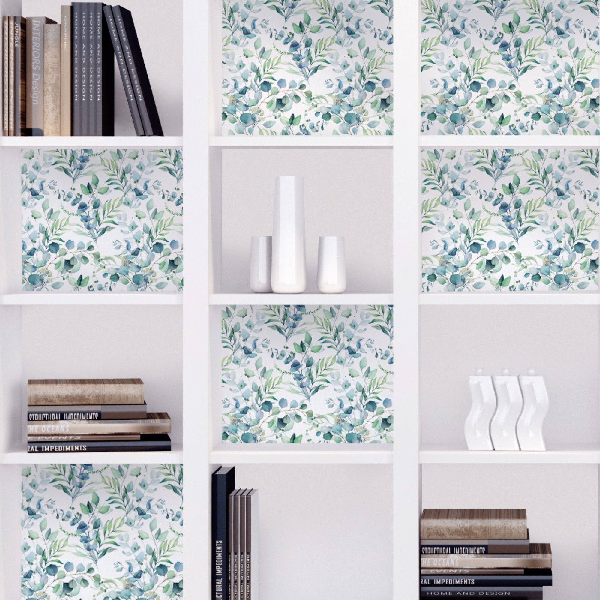 White cube bookcase with eucalyptus-print peel and stick sheets fitted as backing panels inside several cubbies, next to stacked books and white vases