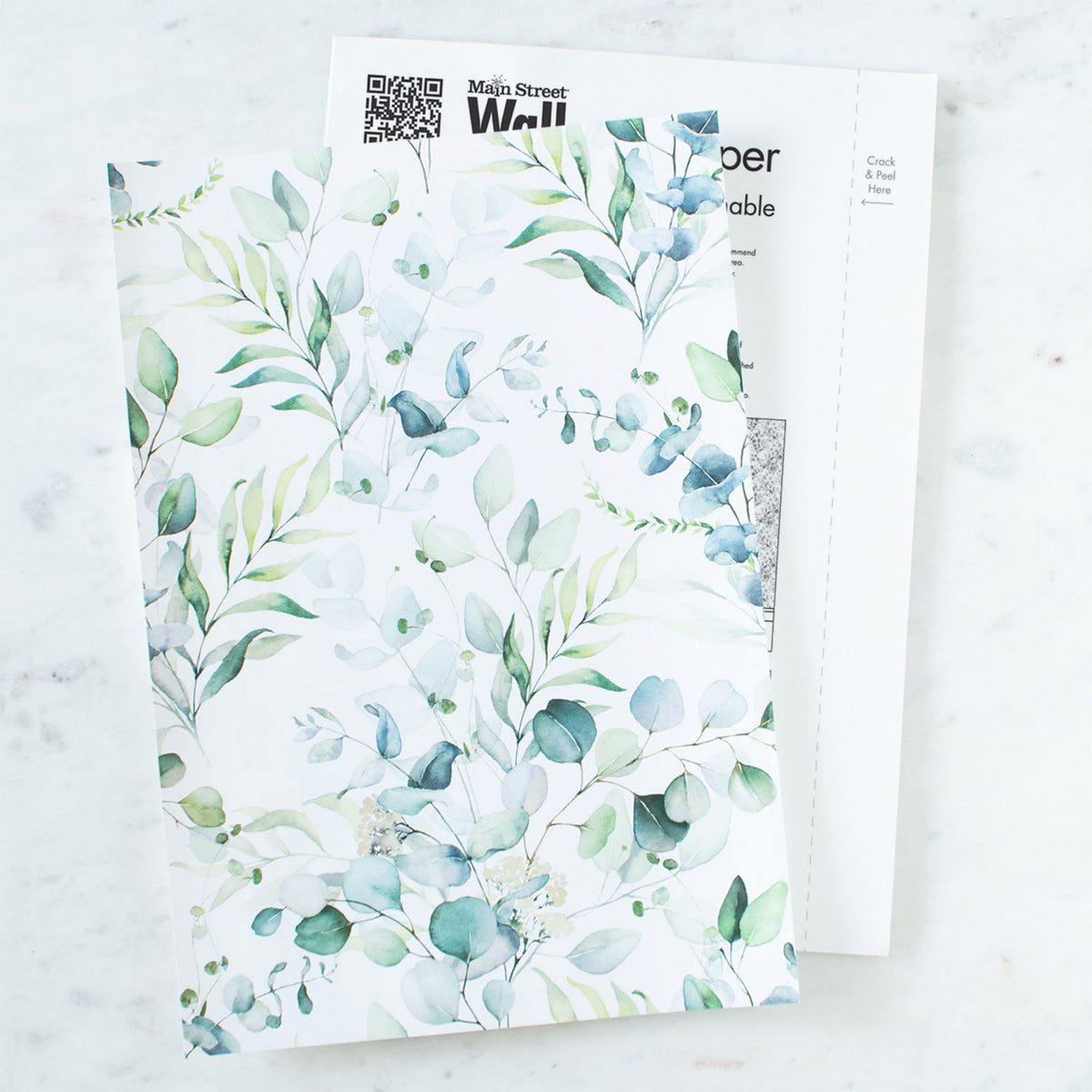 Two Main Street peel and stick wallpaper sheets in a green and blue eucalyptus leaf print resting on a white marble surface, the sheet underneath showing its printed backing with a QR code, the Main Street Wall brand mark and a Crack and Peel Here tab.