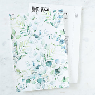 Two Main Street peel and stick wallpaper sheets in a green and blue eucalyptus leaf print resting on a white marble surface, the sheet underneath showing its printed backing with a QR code, the Main Street Wall brand mark and a Crack and Peel Here tab.