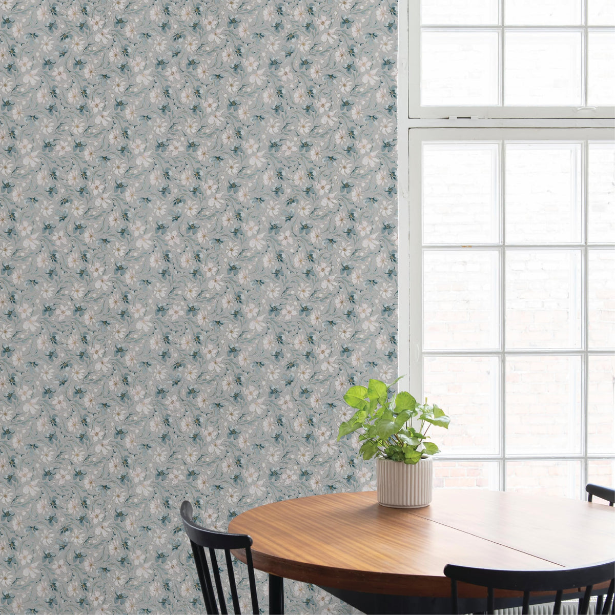 Round wooden dining table with black spindle-back chairs and a potted plant in a ribbed white pot, set against a blue-gray floral wallpapered wall beside a tall paned window