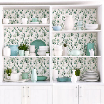 A white built-in hutch with open shelves whose back panels are covered in a green and white leaf print, holding white and sage-green pitchers, plates, bowls, cups and small potted plants above closed white cabinet doors.