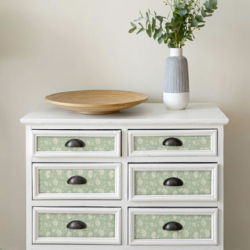 A white six-drawer dresser with sage green floral peel and stick paper set into each drawer front panel, holding a shallow wooden bowl and a grey striped vase of eucalyptus.