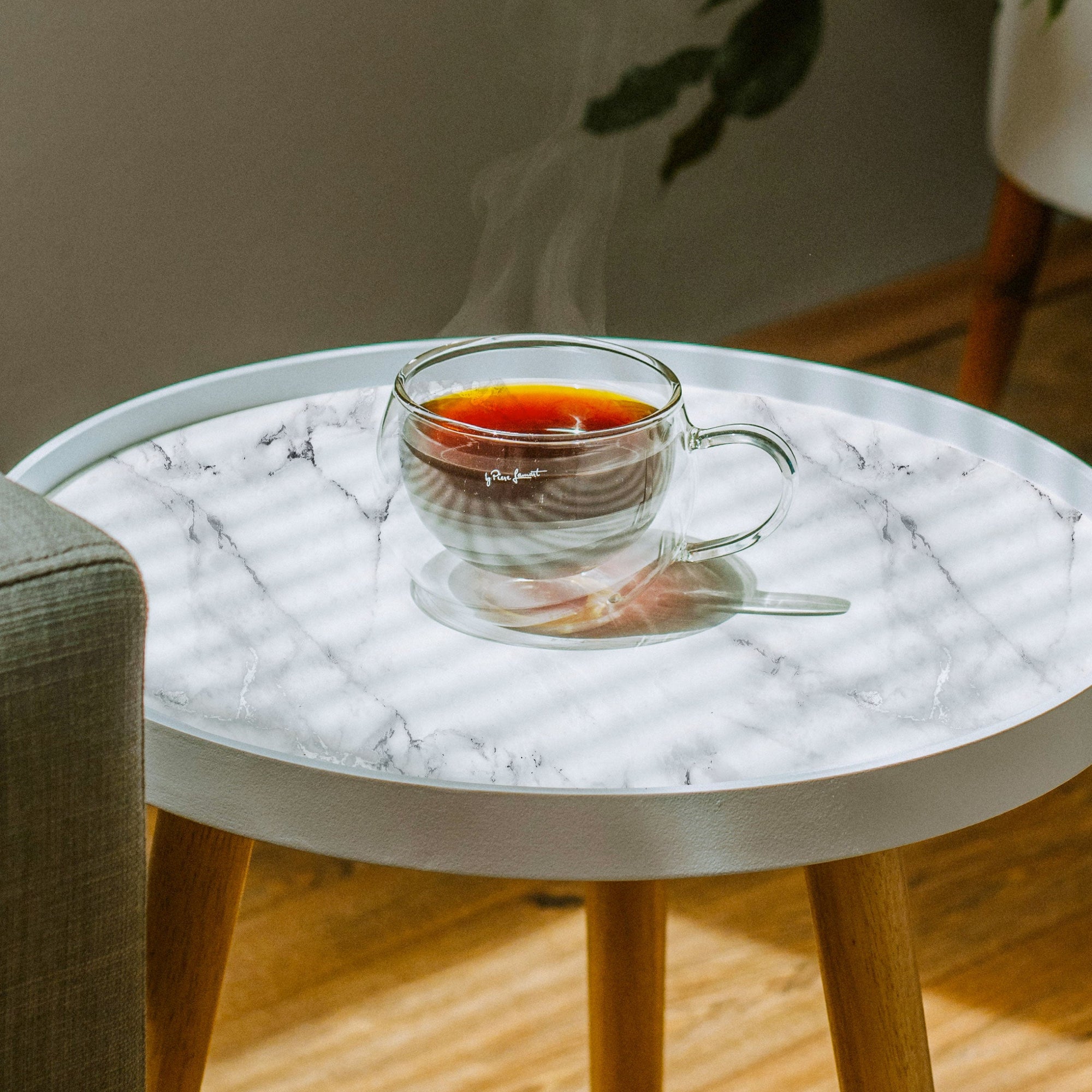 A round white side table topped with white marble-pattern peel and stick paper, holding a clear glass cup of black tea in a sunlit room.