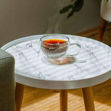 A round white side table topped with white marble-pattern peel and stick paper, holding a clear glass cup of black tea in a sunlit room.