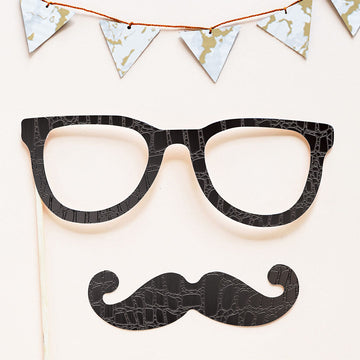 Photo booth props cut from black embossed craft paper, a pair of glasses on a wooden stick and a curled mustache, below a string of white and gold marbled paper triangles