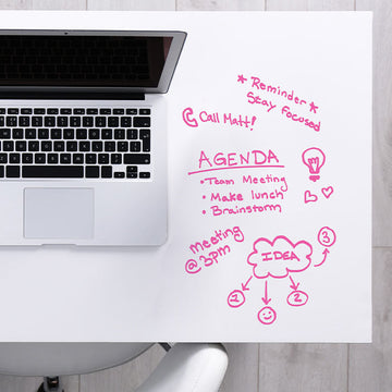 White desk surface covered in dry erase paper and filled with pink marker notes and an agenda list beside an open laptop.