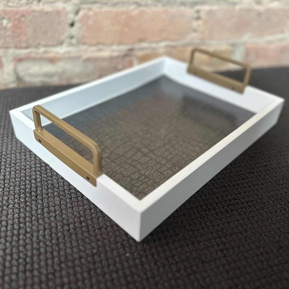 A white wooden serving tray with gold metal handles, its base lined with dark grey crocodile-embossed paper, set on a dark woven mat in front of a brick wall.