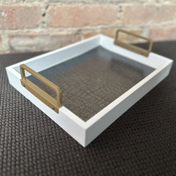 A white wooden serving tray with gold metal handles, its base lined with dark grey crocodile-embossed paper, set on a dark woven mat in front of a brick wall.