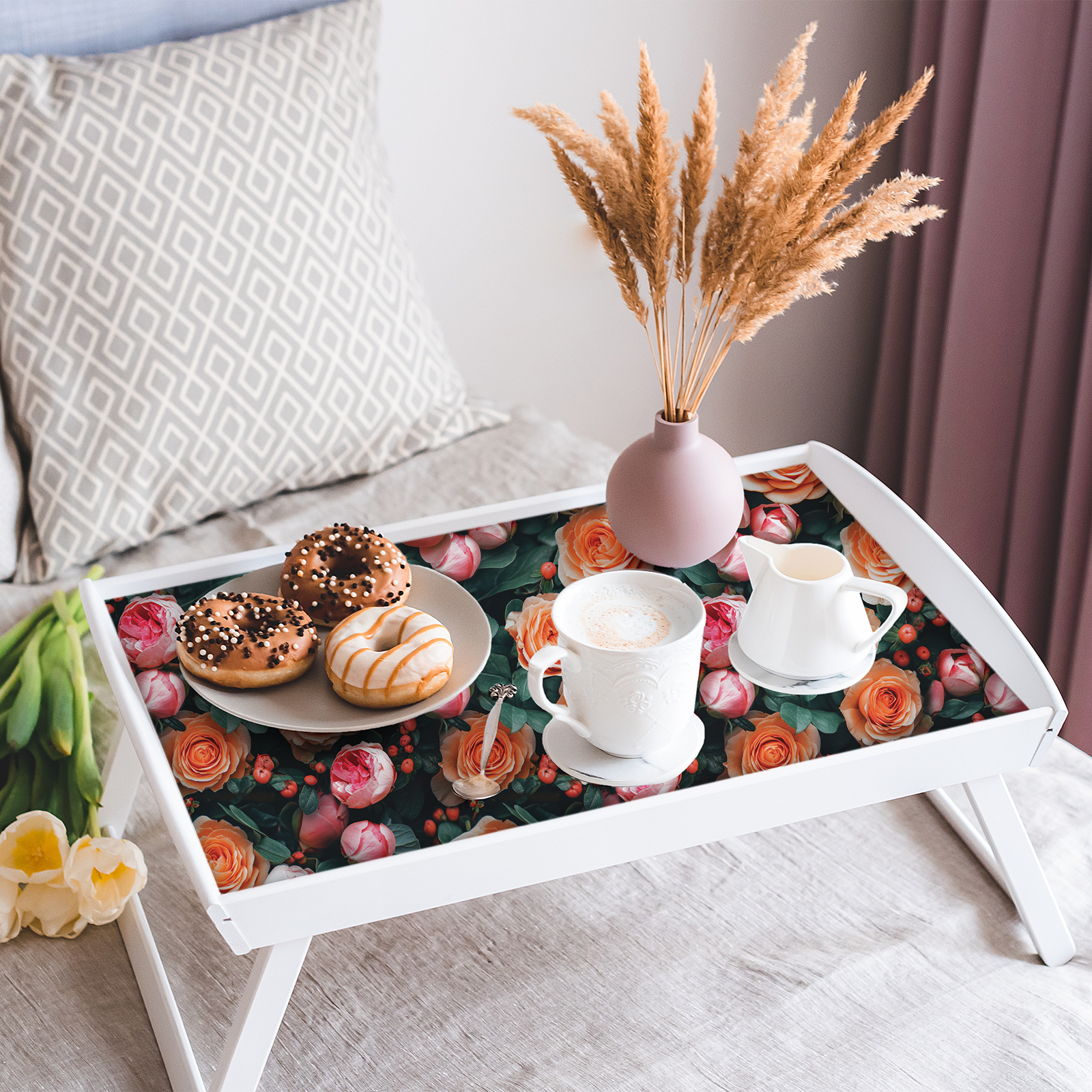 A white folding bed tray with its flat base lined in Fresh Roses peel and stick paper, set with a plate of donuts, a cappuccino, a creamer and a pink vase of dried pampas grass.