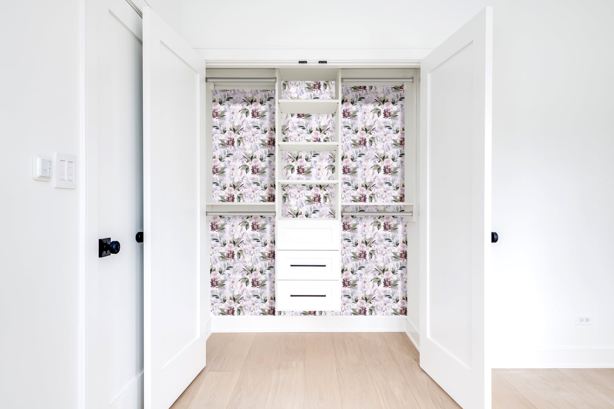 A white reach-in closet with the back wall and shelf faces papered in a soft lilac floral print, hanging rods on both sides and a stack of three white drawers at the bottom.