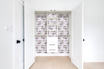 A white reach-in closet with the back wall and shelf faces papered in a soft lilac floral print, hanging rods on both sides and a stack of three white drawers at the bottom.
