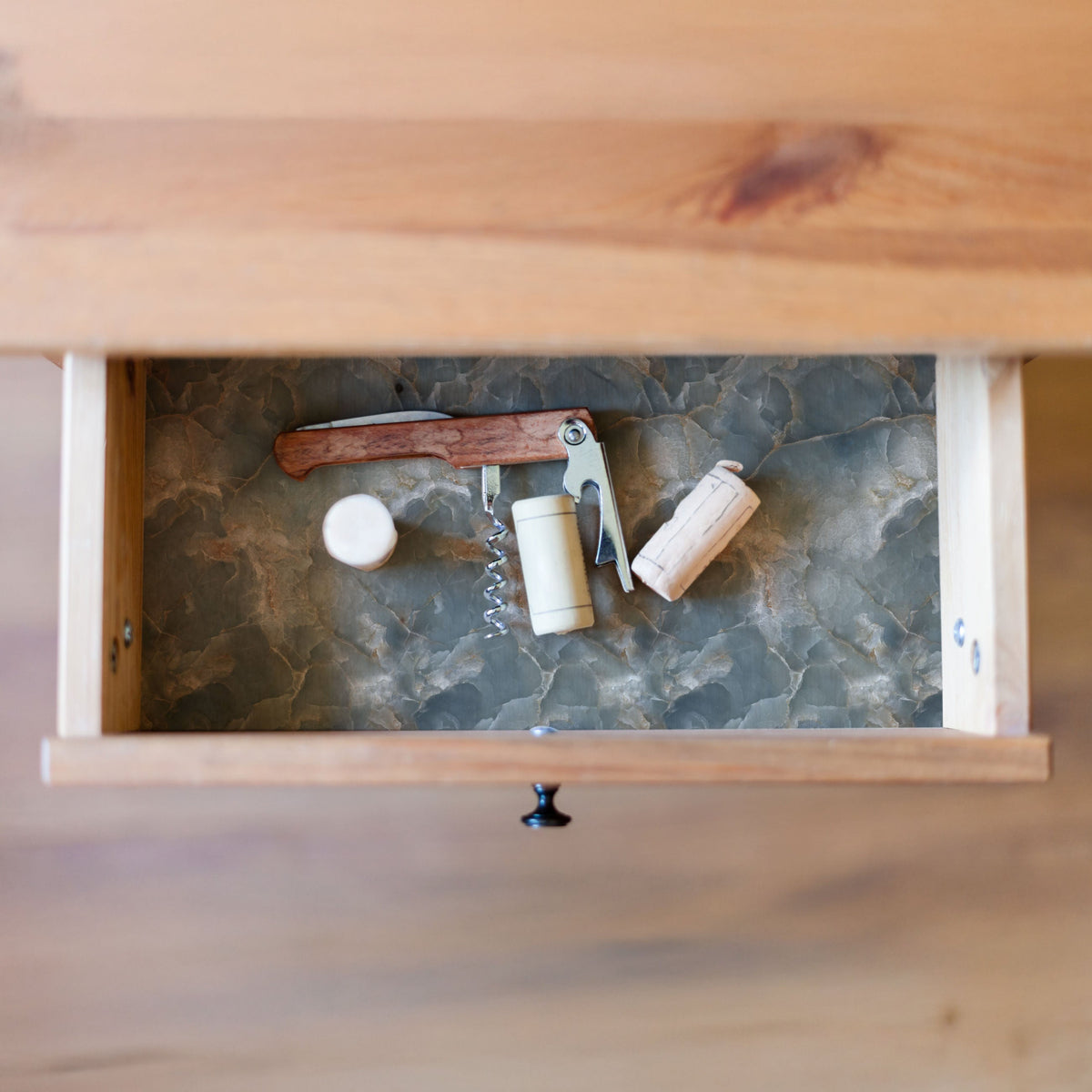Overhead view of an open wooden drawer lined with gray-green stone-look paper, holding a waiter's corkscrew with a wooden handle and three corks