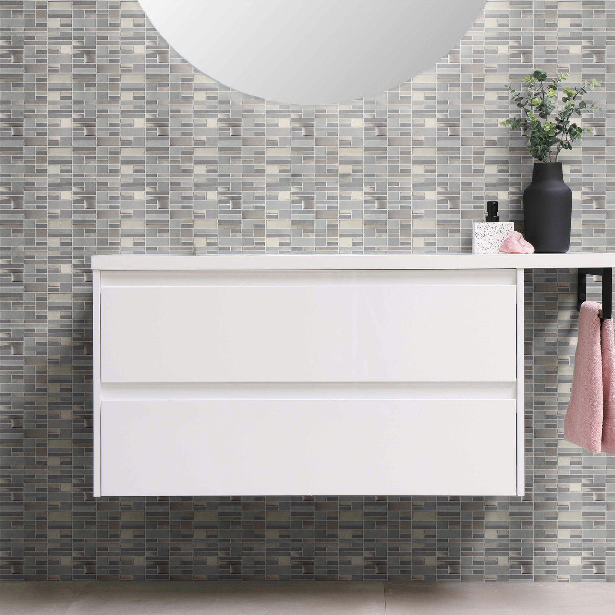 White two-drawer floating bathroom vanity with a round mirror, a black vase of greenery and a pink hand towel, against a wall of gray and silver mosaic-look peel and stick wallpaper tiles