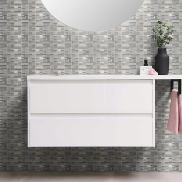 White two-drawer floating bathroom vanity with a round mirror, a black vase of greenery and a pink hand towel, against a wall of gray and silver mosaic-look peel and stick wallpaper tiles