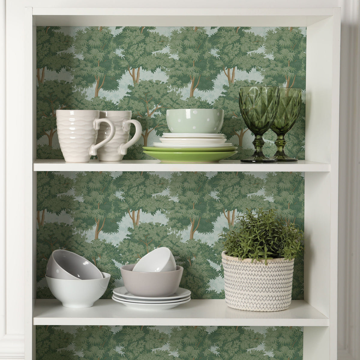 White open shelving unit lined along the back with a green tree print, holding white mugs, stacked plates and bowls, green glass goblets and a potted plant
