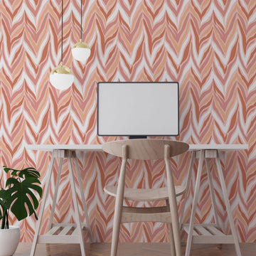 A home office wall covered in coral and blush chevron peel and stick wallpaper, behind a white trestle desk, wooden chair, two brass pendant lights and a potted monstera.
